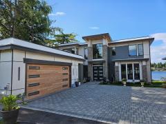 Front door & Garage Modern two-story house with large windows, a flat roof, and a wooden double garage door, situated by a lake under a clear blue sky.