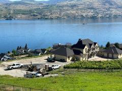 Lake Chelan Residence from above Large house under construction near a lake, with construction vehicles and materials in the driveway; hills and water are visible in the background.